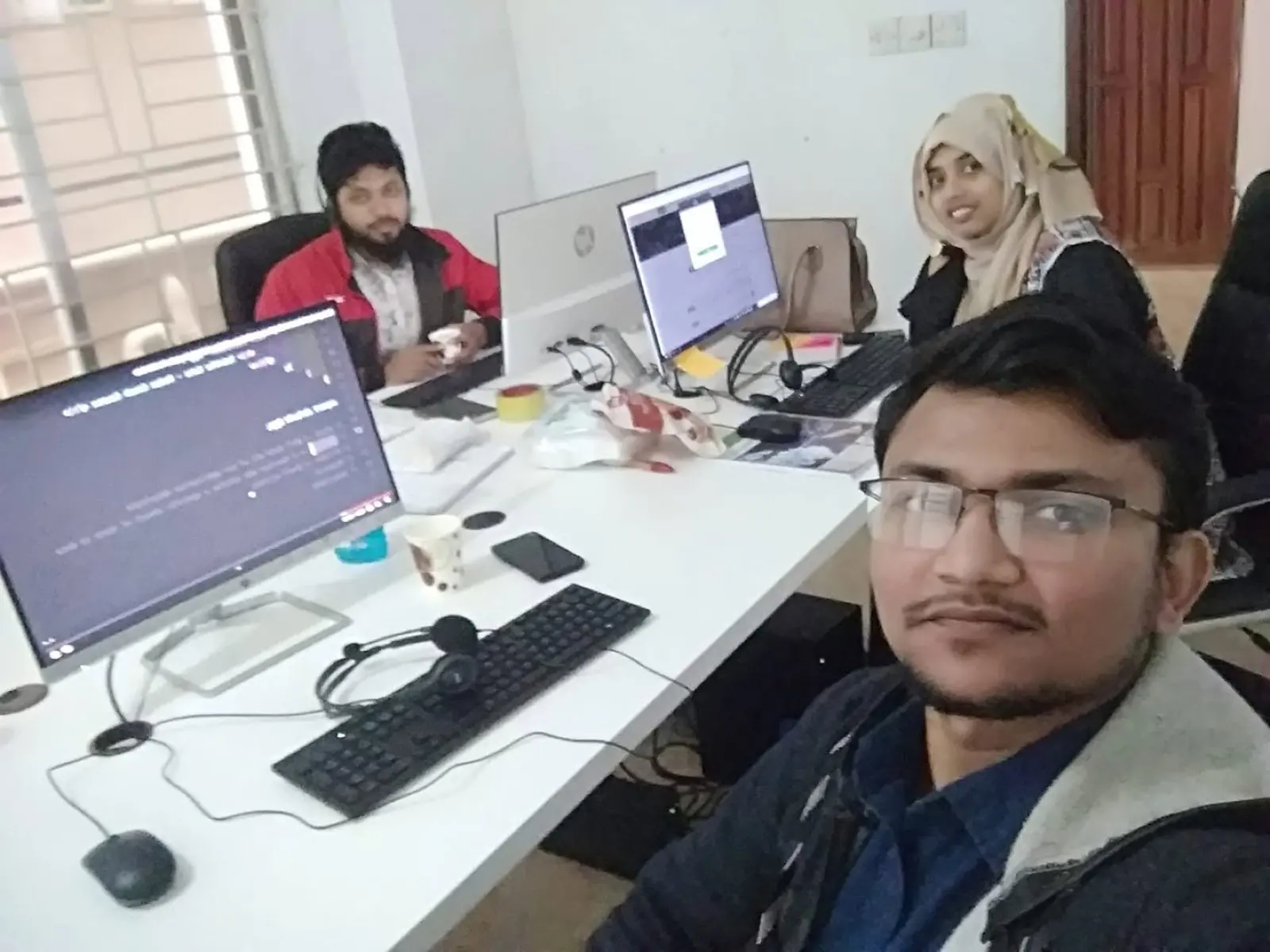 Three developers at a workstation with multiple monitors, source code visible on the nearest screen