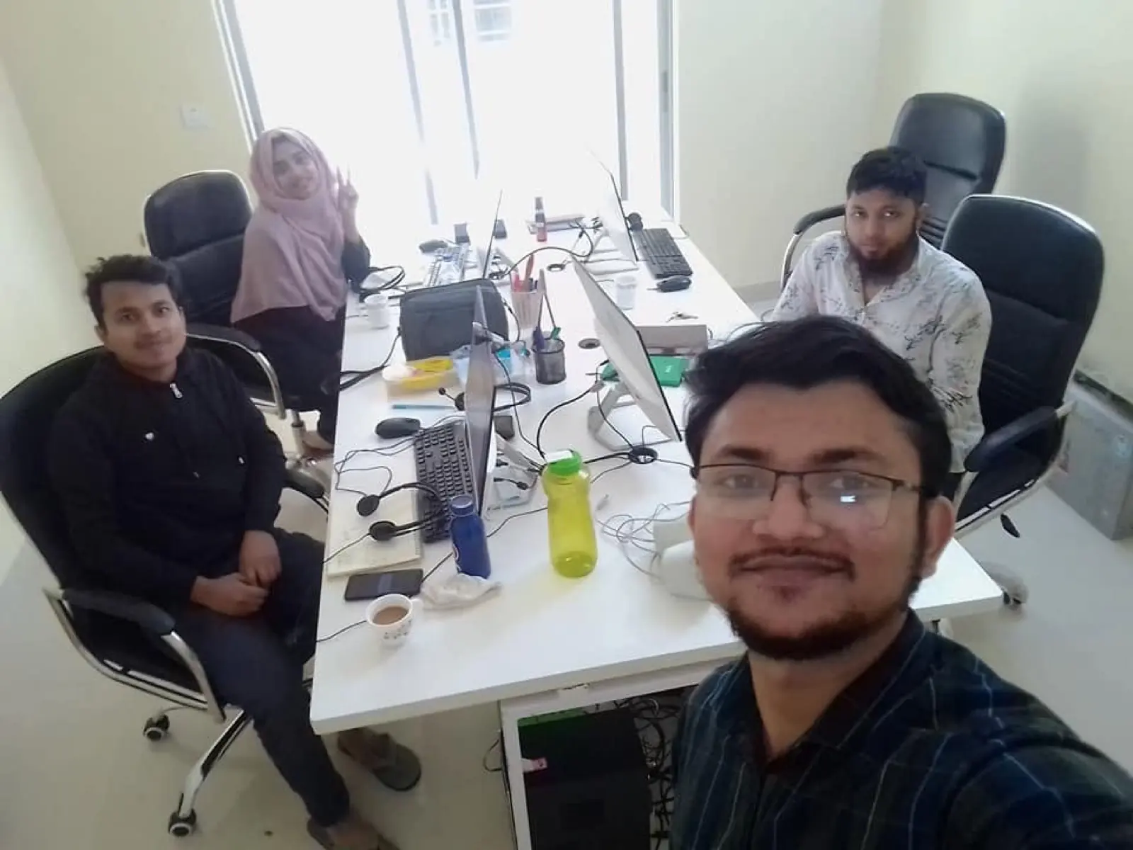 Colleagues seated along a long shared desk working on laptops and monitors in a bright, sunlit office