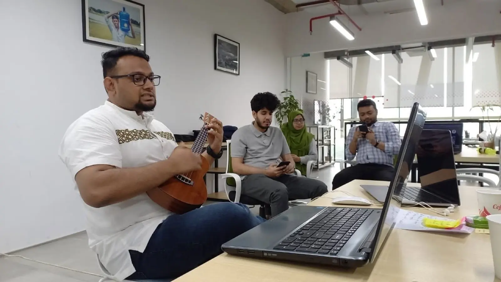 A colleague playing a small guitar at his desk while three teammates sit nearby in the office